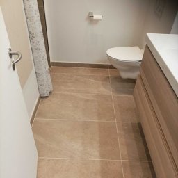 Karol Piątkowski - Wykończona łazienka z beżowymi płytkami podłogowymi, wiszącą toaletą, umywalką z szafką z jasnego drewna i zasłoną prysznicową w kwiaty.