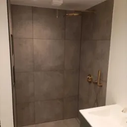 Nowoczesna kabina prysznicowa z dużymi, szarymi płytkami imitującymi beton, deszczownicą w kolorze mosiądzu i minimalistyczną armaturą, widoczna umywalka z blatem.