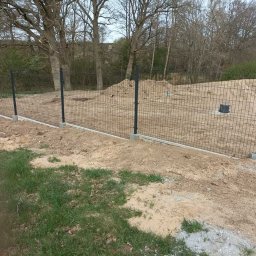 Karol Piątkowski - Nowo postawione, metalowe ogrodzenie panelowe z podmurówką betonową na nierównym terenie, widok na drzewa i hałdę ziemi w tle.