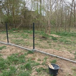 Karol Piątkowski - Montaż ogrodzenia panelowego z podmurówką betonową na działce z widocznym wiaderkiem z drewnianymi kołkami.