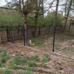 Karol Piątkowski - Nowo postawione, metalowe ogrodzenie panelowe w trakcie montażu na betonowej podmurówce, widoczne drzewa i drewniany płot w tle oraz fragment strzechy budynku.