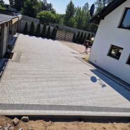 Karol Piątkowski - Nowo ułożona, jasnoszara kostka brukowa na podjeździe przed domem, z widocznym fragmentem ogrodu i osobą myjącą powierzchnię w tle.