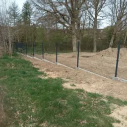 Nowo postawione, czarne ogrodzenie panelowe z betonową podmurówką na granicy działki z młodym lasem, widok z perspektywy trawy.