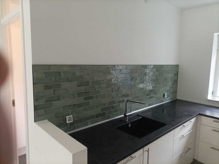 Kuchnia z zieloną, ceglaną mozaiką nad czarnym blatem i białymi szafkami, widoczny kran i okno.