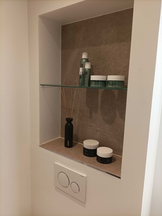 Wnęka w łazience wyłożona brązowymi płytkami, z szklaną półką, na której stoją kosmetyki w zielonych opakowaniach i czarny dyfuzor zapachowy, poniżej dwa małe słoiczki z kremami, a obok przycisk...