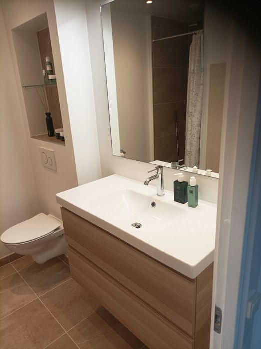 Nowoczesna łazienka z umywalką nablatową na drewnianej szafce, lustrem, wnęką z dekoracjami, wiszącą toaletą i brązowymi płytkami podłogowymi.