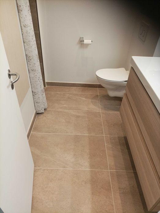 Wykończona łazienka z beżowymi płytkami podłogowymi, wiszącą toaletą, umywalką z szafką z jasnego drewna i zasłoną prysznicową w kwiaty.