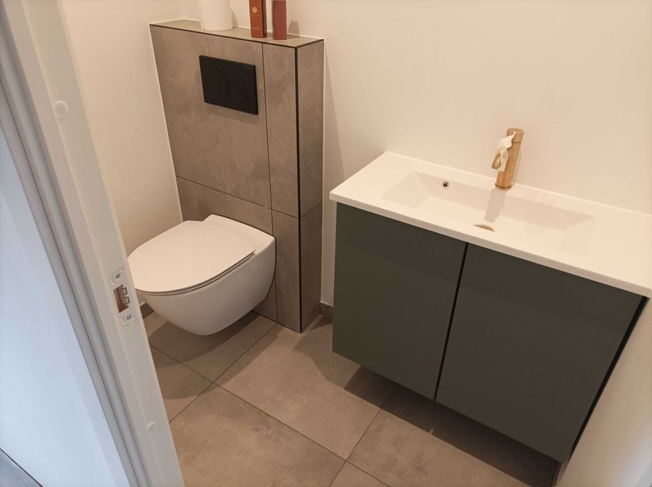 Nowoczesna łazienka z podwieszaną toaletą, zabudową z płytek imitujących kamień i szafką pod umywalką w kolorze oliwkowym, wykończona złotą armaturą.