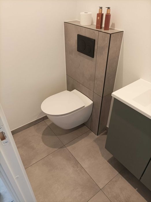 Nowoczesna łazienka z podwieszaną toaletą, szarą zabudową z czarnym przyciskiem spłukiwania, beżowymi płytkami podłogowymi i kosmetykami na górze zabudowy.