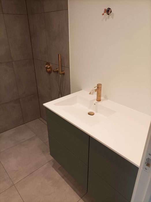 Nowoczesna łazienka z szarymi płytkami, złotą armaturą i umywalką z szafką w odcieniach zieleni, widoczne przyłącze instalacji na ścianie.