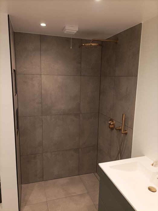 Nowoczesna kabina prysznicowa z dużymi, szarymi płytkami imitującymi beton, deszczownicą w kolorze mosiądzu i minimalistyczną armaturą, widoczna umywalka z blatem.