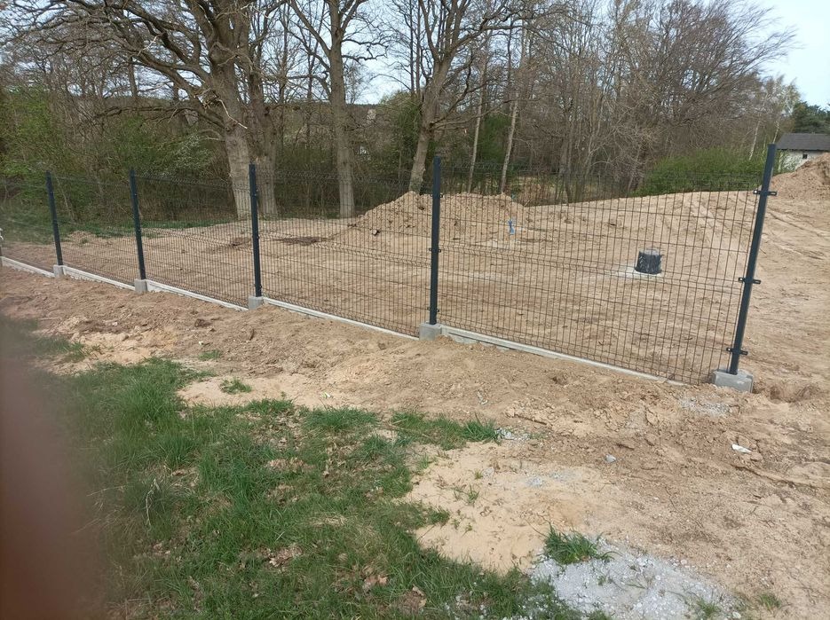 Nowo postawione, metalowe ogrodzenie panelowe z podmurówką betonową na nierównym terenie, widok na drzewa i hałdę ziemi w tle.