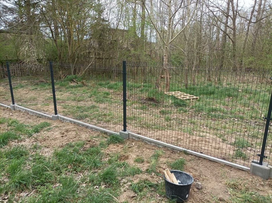 Montaż ogrodzenia panelowego z podmurówką betonową na działce z widocznym wiaderkiem z drewnianymi kołkami.