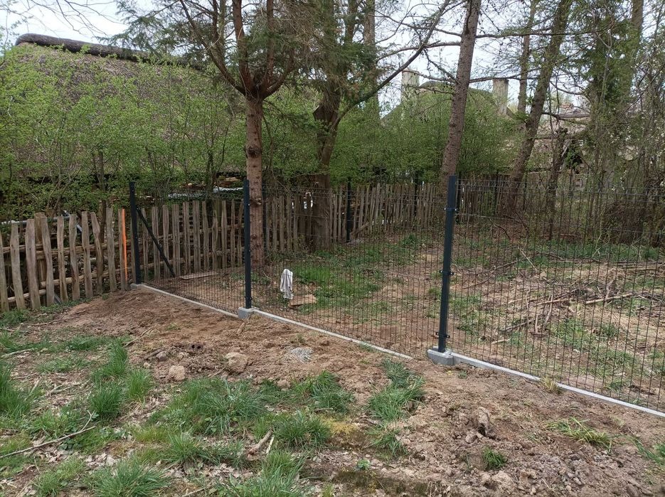 Nowo postawione, metalowe ogrodzenie panelowe w trakcie montażu na betonowej podmurówce, widoczne drzewa i drewniany płot w tle oraz fragment strzechy budynku.