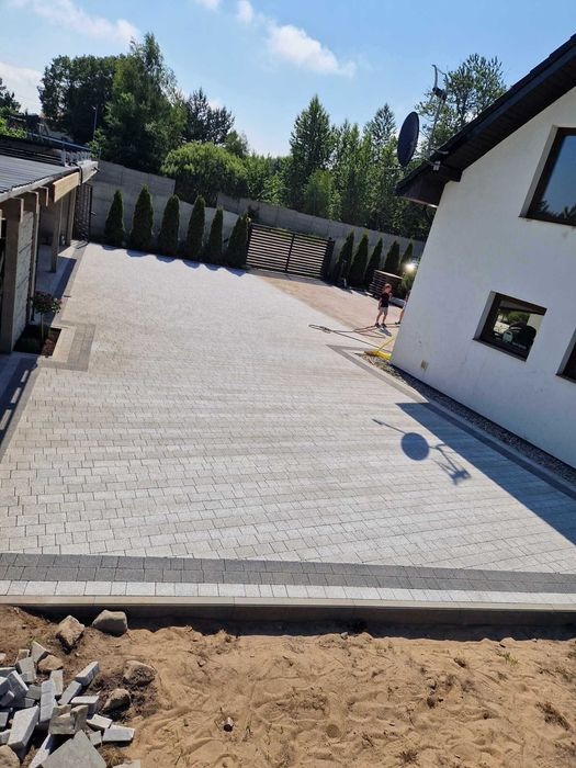 Nowo ułożona, jasnoszara kostka brukowa na podjeździe przed domem, z widocznym fragmentem ogrodu i osobą myjącą powierzchnię w tle.