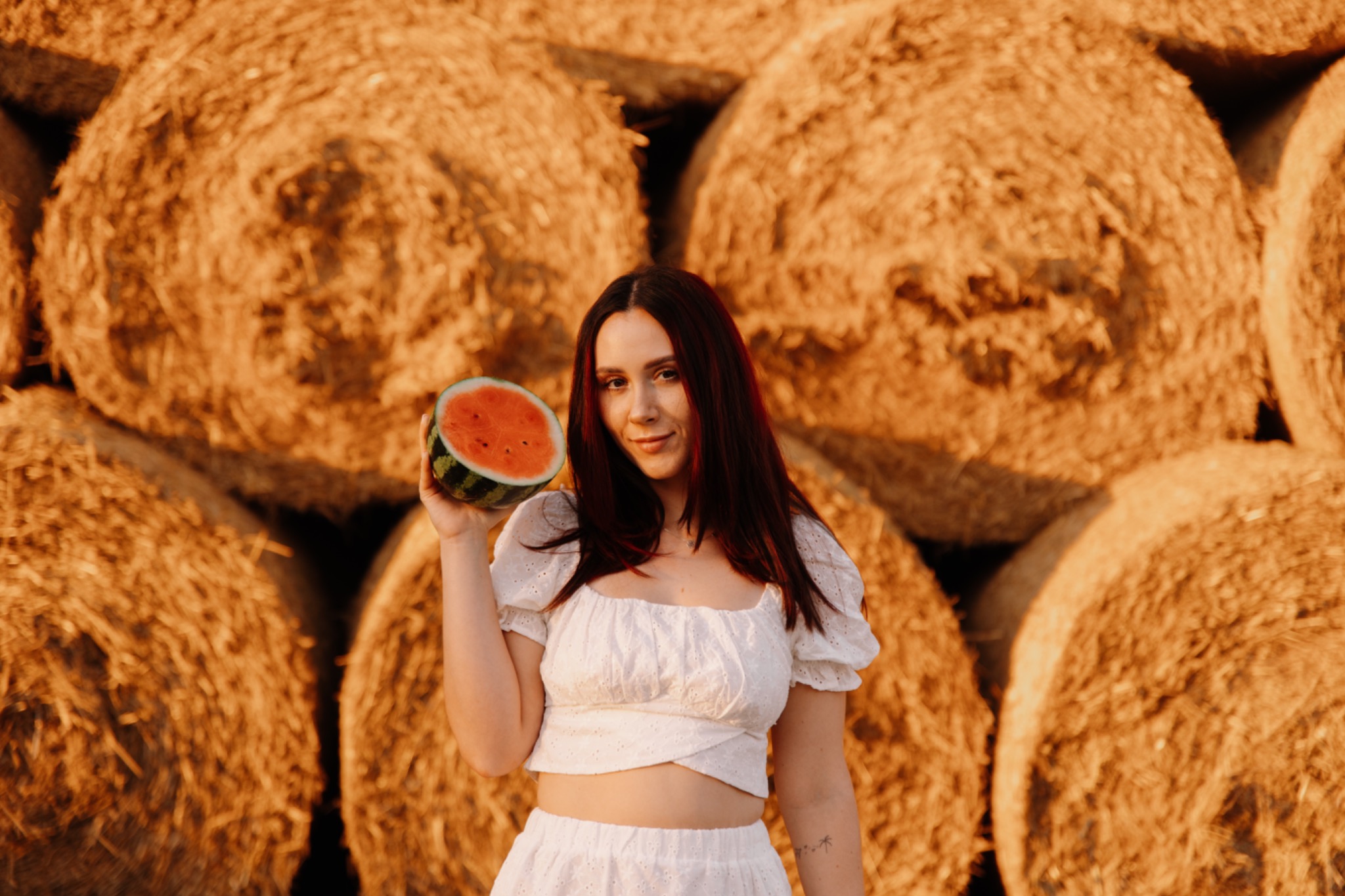 Uśmiechnięta kobieta w białym stroju trzymająca przekrojonego arbuza na tle stogów siana w ciepłym, zachodzącym słońcu.