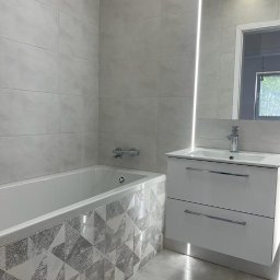 VM-BUD - Nowoczesna łazienka z wanną obudowaną panelami w geometryczne wzory, szare płytki na ścianach, biała szafka pod umywalką z chromowaną baterią, podświetlane lustro.