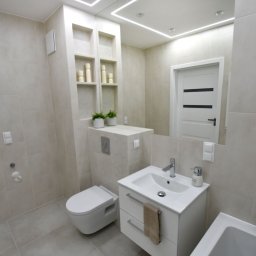 VM-BUD - Jasna łazienka z beżowymi płytkami, podwieszana toaleta, biała szafka pod umywalką z szufladami i ręcznikiem, lustro z oświetleniem LED, oraz wnęka z półkami i dekoracjami.