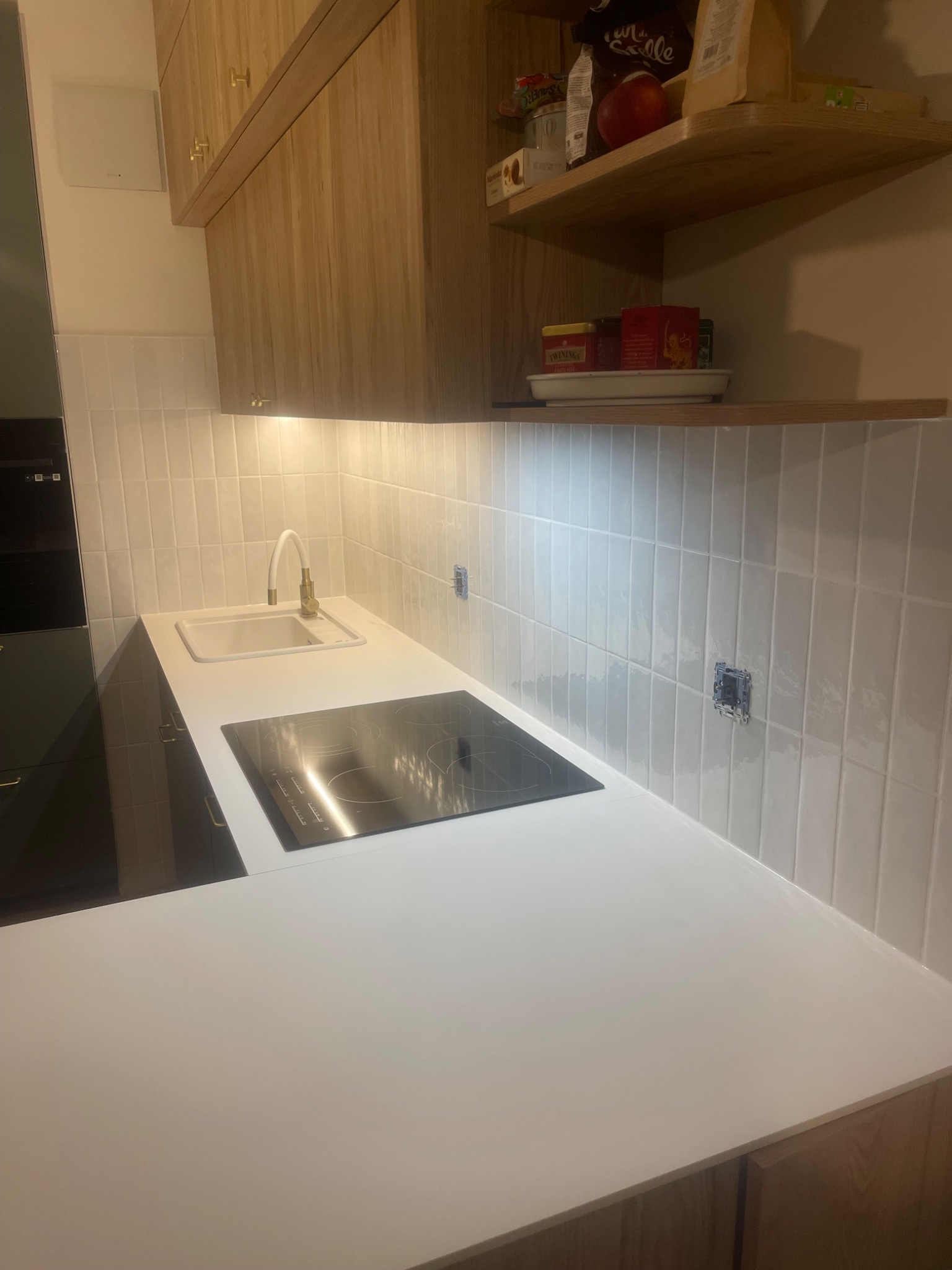 Nowoczesna kuchnia po remoncie: białe, pionowo ułożone płytki na ścianie, biały blat, złota bateria przy zlewie i płyta indukcyjna. Widoczne niedokończone gniazdko elektryczne.