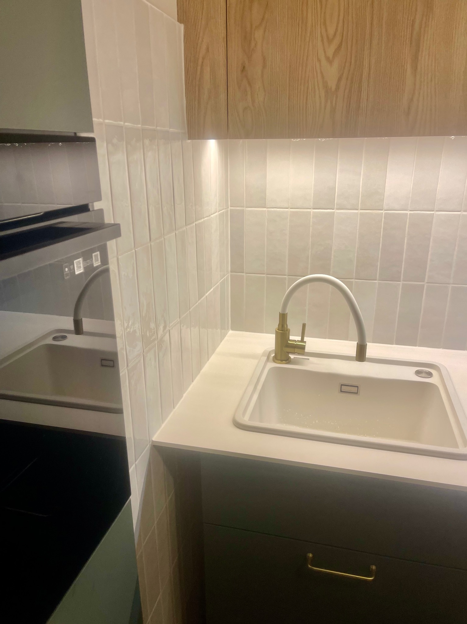 Nowoczesna kuchnia z białymi, pionowo ułożonymi płytkami, złotą baterią przy białym zlewie i drewnianą szafką wiszącą.