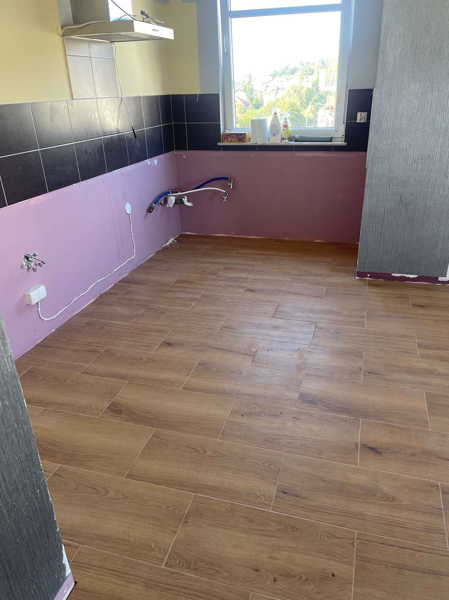 Kuchnia w trakcie prac wykończeniowych: położona podłoga z paneli imitujących drewno, ściany przygotowane pod płytki w kolorze fioletowym, widoczne instalacje wodne i elektryczne.
