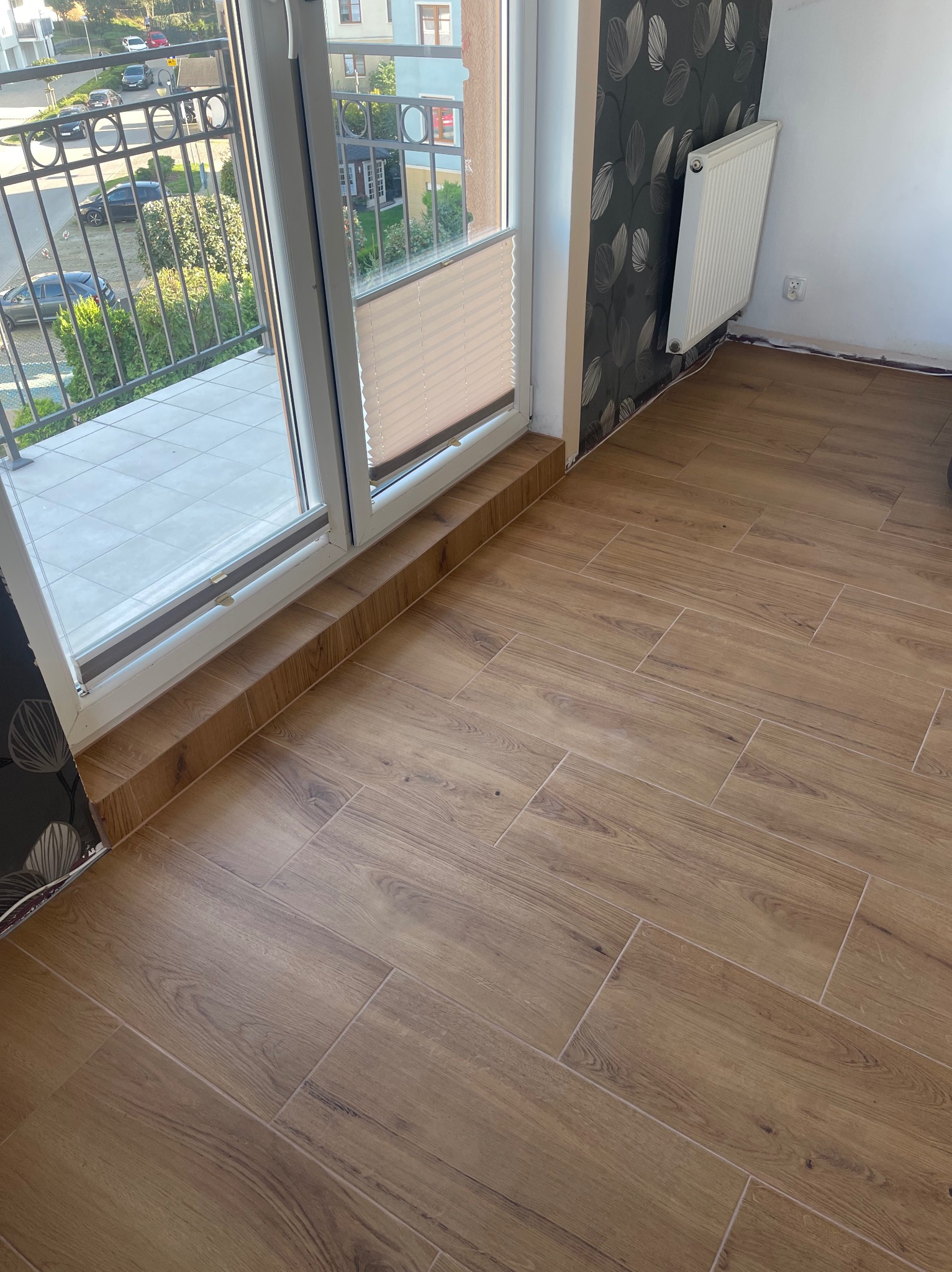 Ułożona podłoga z płytek imitujących drewno w pokoju z balkonem, widoczny grzejnik i fragment wzorzystej tapety.