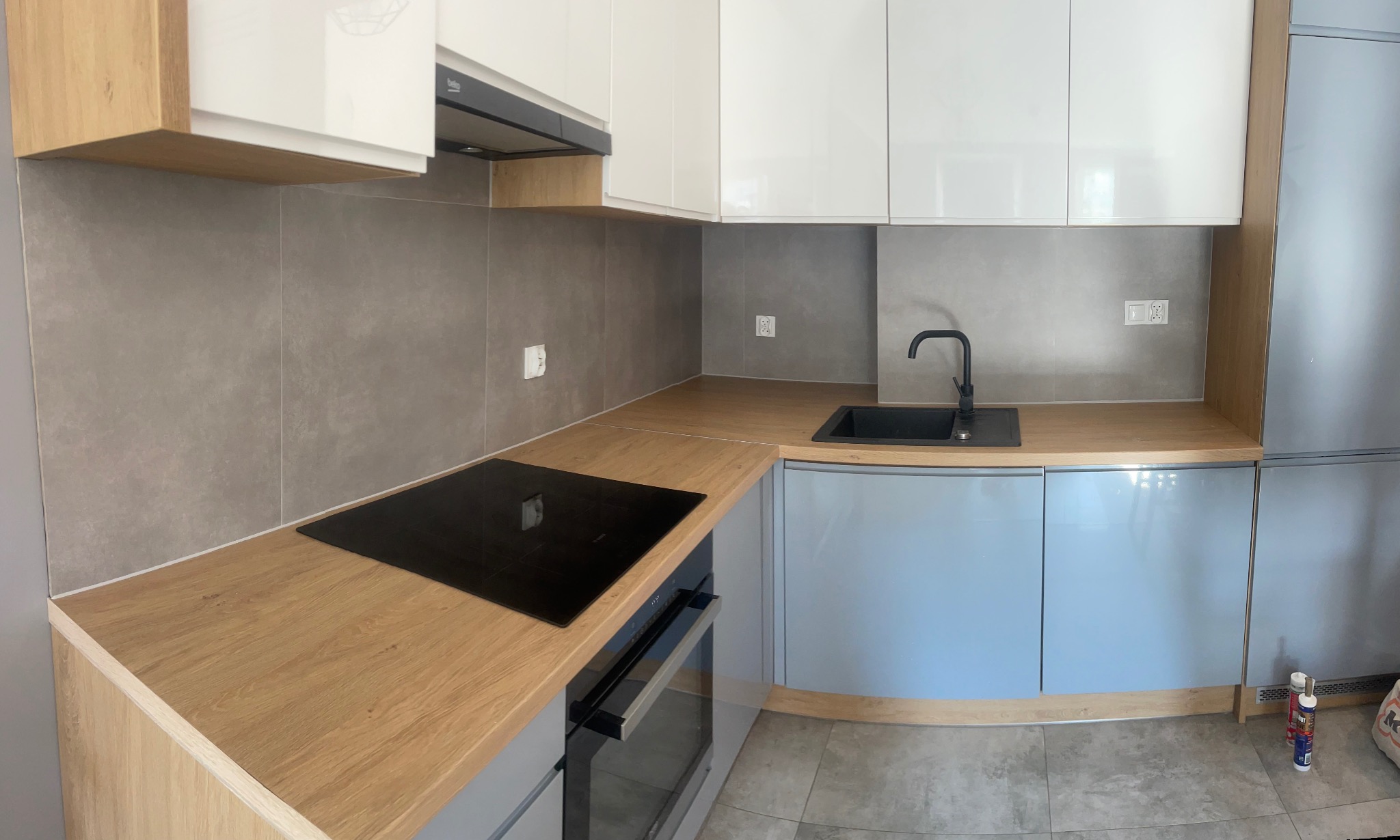 Nowoczesna kuchnia po remoncie: szare płytki ścienne, drewniany blat, białe i szare szafki, czarna płyta indukcyjna i zlew, widoczne gniazdka elektryczne.