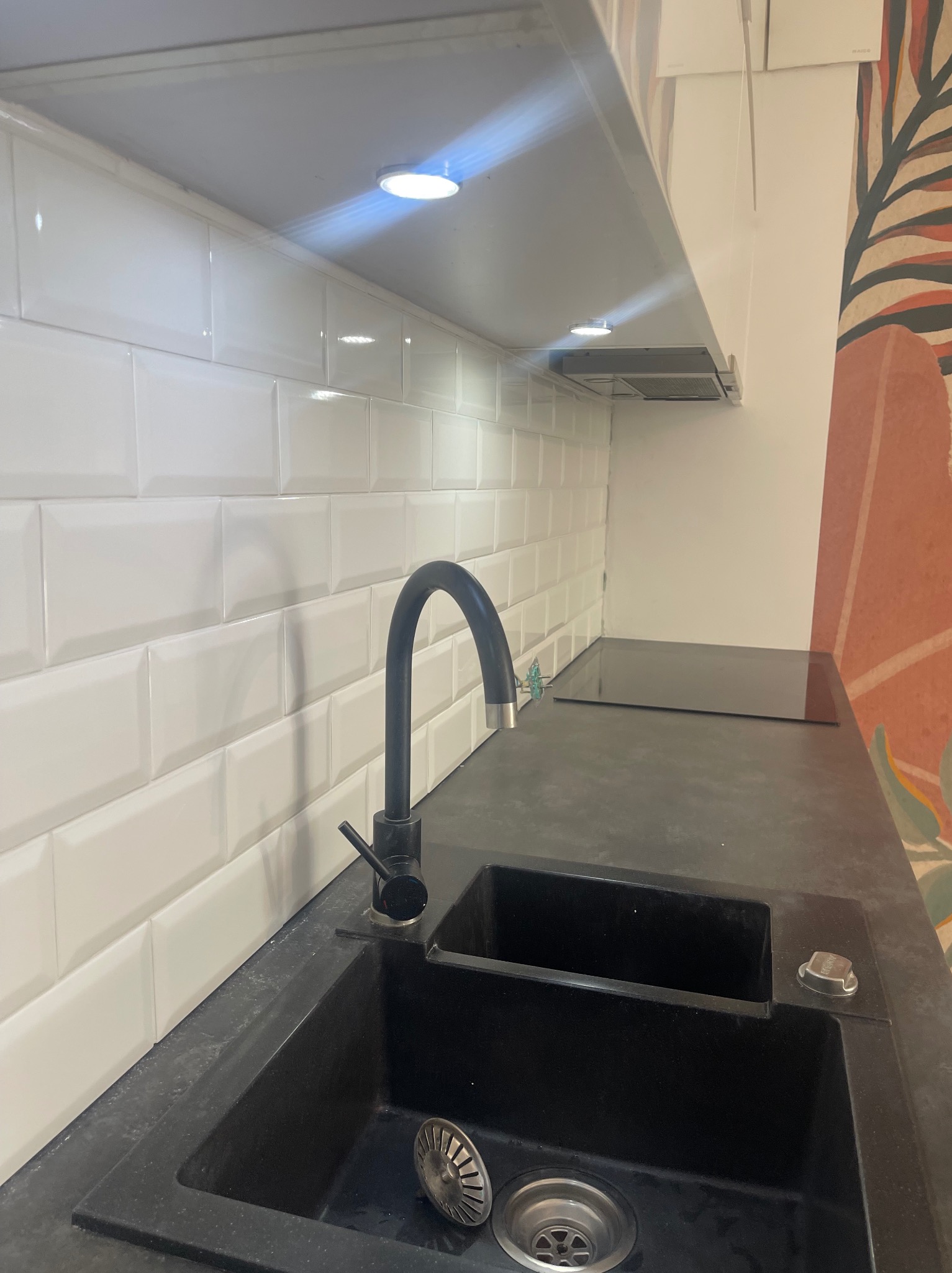 Czarny, dwukomorowy zlew z czarną baterią i metalowym sitkiem w komorze, białe, błyszczące płytki na ścianie, fragment dekoracyjnej tapety z motywem roślinnym w tle.