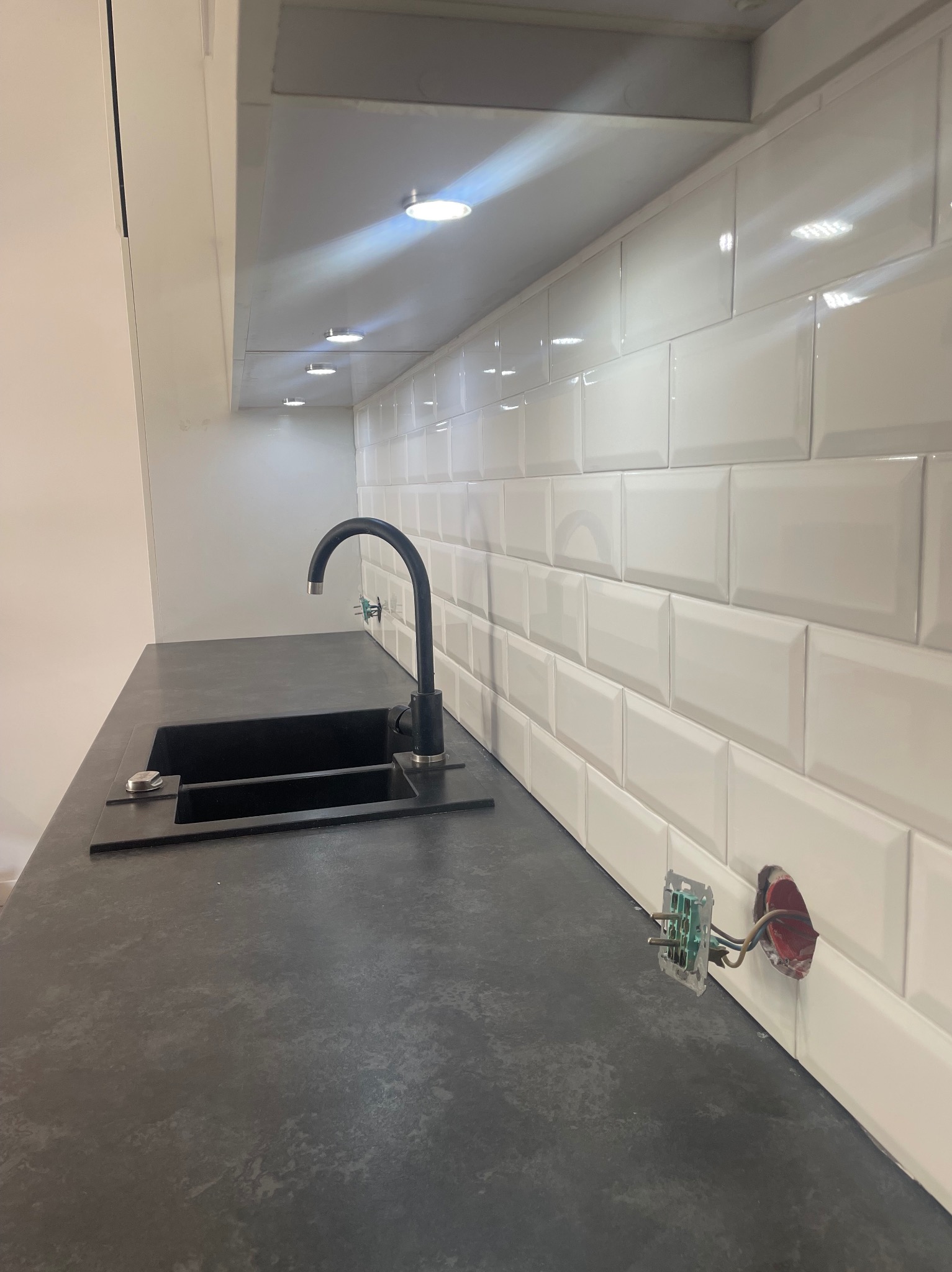 Częściowo wykończona kuchnia: czarny zlew i bateria na blacie w kolorze ciemnego grafitu, białe płytki typu metro na ścianie, widoczne przewody elektryczne w puszce.