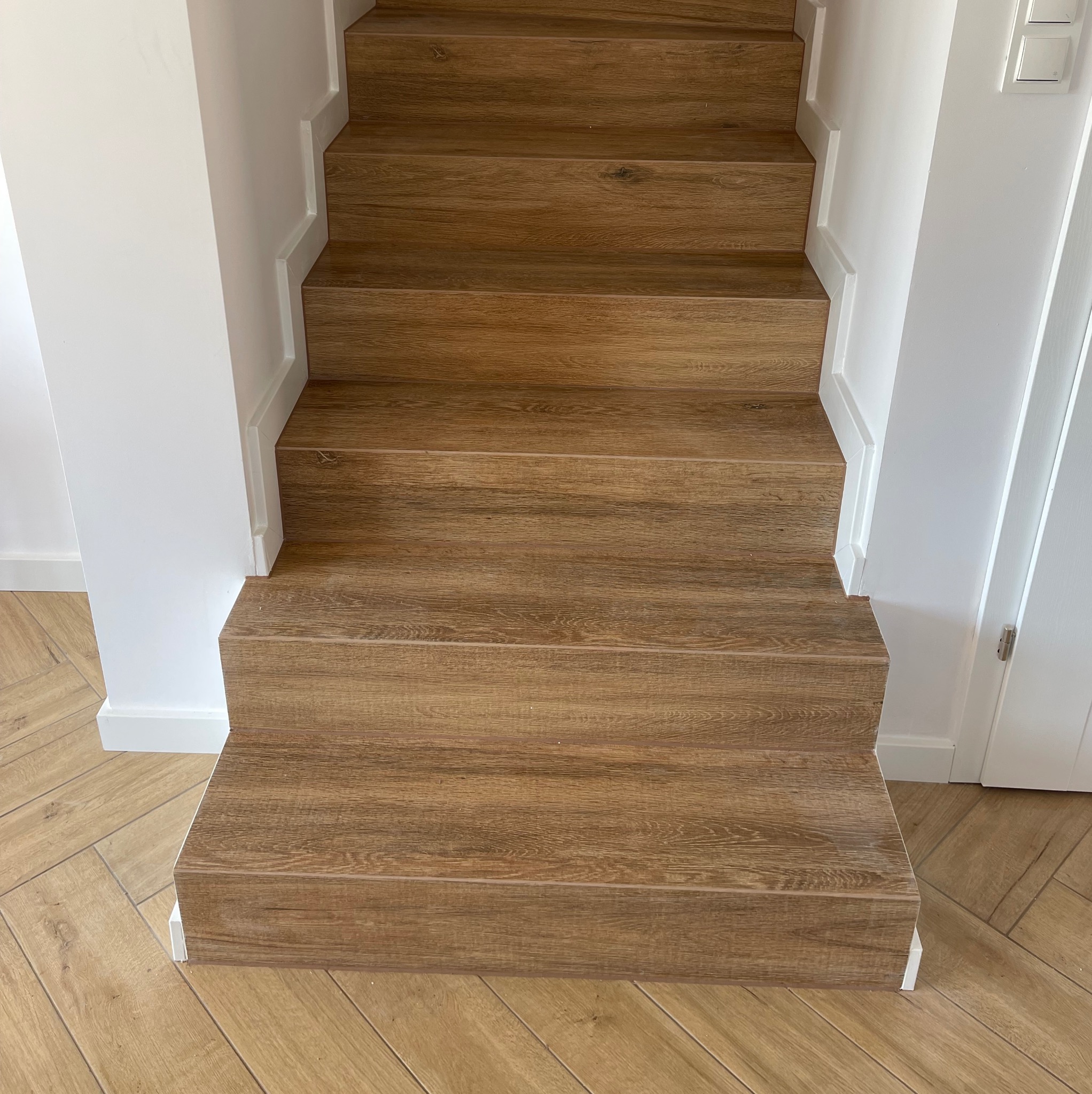 Schody wyłożone panelami imitującymi drewno, z białymi listwami przypodłogowymi i białymi ścianami, podłoga wyłożona parkietem w jodełkę.