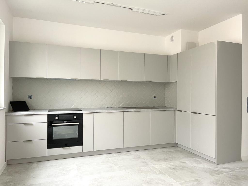 Jasnoszara, minimalistyczna kuchnia z płytkami ułożonymi w jodełkę na ścianie, czarnym piekarnikiem Amica i szarymi płytkami podłogowymi imitującymi kamień.