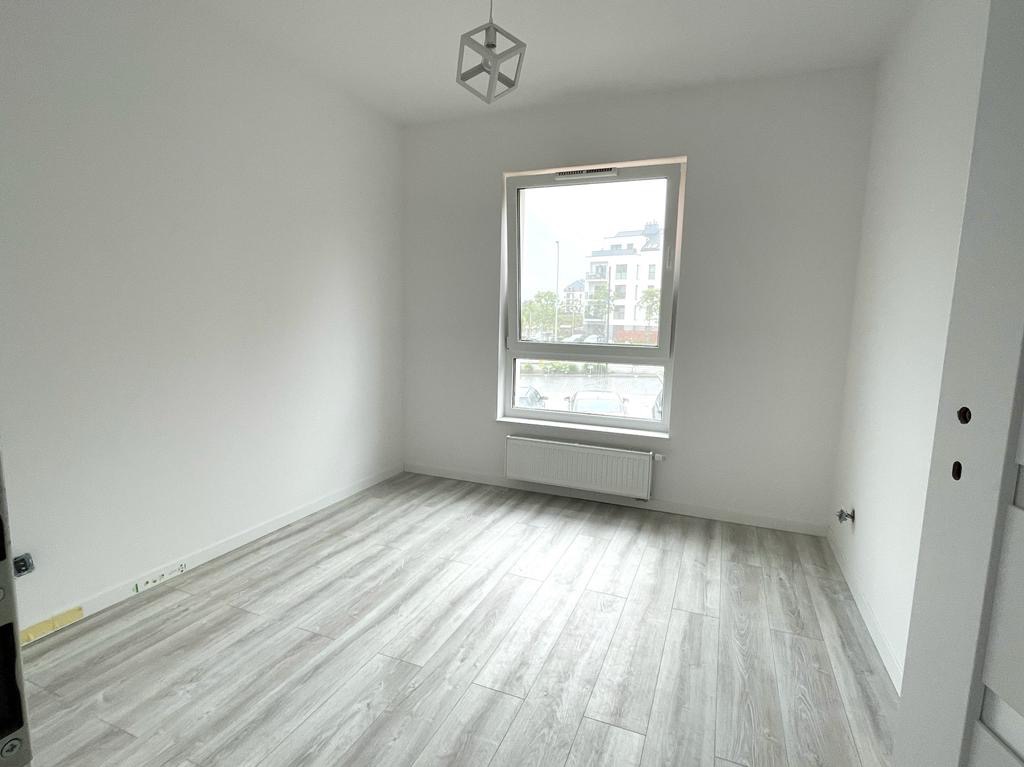 Pusty pokój z panelami podłogowymi imitującymi drewno, białe ściany, okno z widokiem na budynek mieszkalny i geometryczna lampa wisząca.