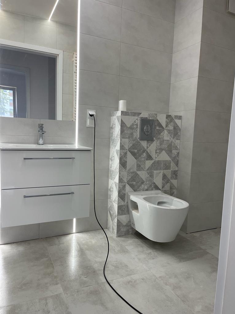 Nowoczesna łazienka z szarymi płytkami, podwieszana toaleta z geometrycznym wzorem na obudowie, biała szafka pod umywalką z dwoma szufladami i podświetlanym lustrem.