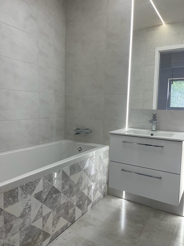 Nowoczesna łazienka z wanną obudowaną panelami w geometryczne wzory, szare płytki na ścianach, biała szafka pod umywalką z chromowaną baterią, podświetlane lustro.