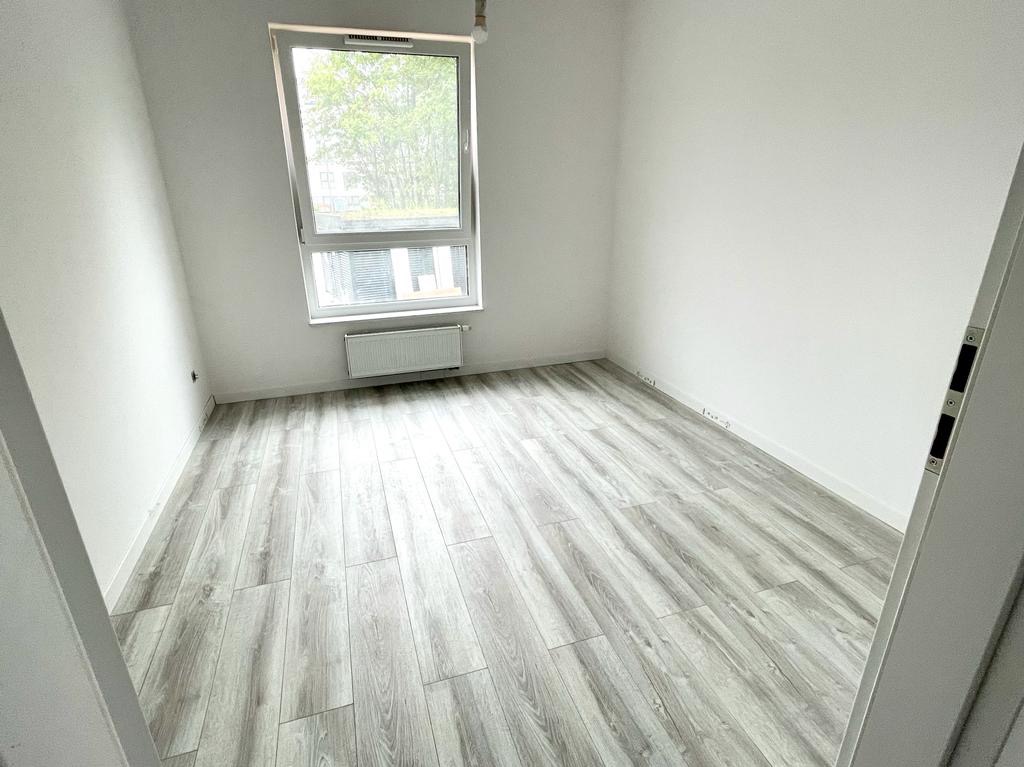 Pusty pokój z ułożonymi panelami podłogowymi w odcieniach szarości, widok na okno z grzejnikiem, białe ściany, widoczny fragment drzwi z prawej strony.