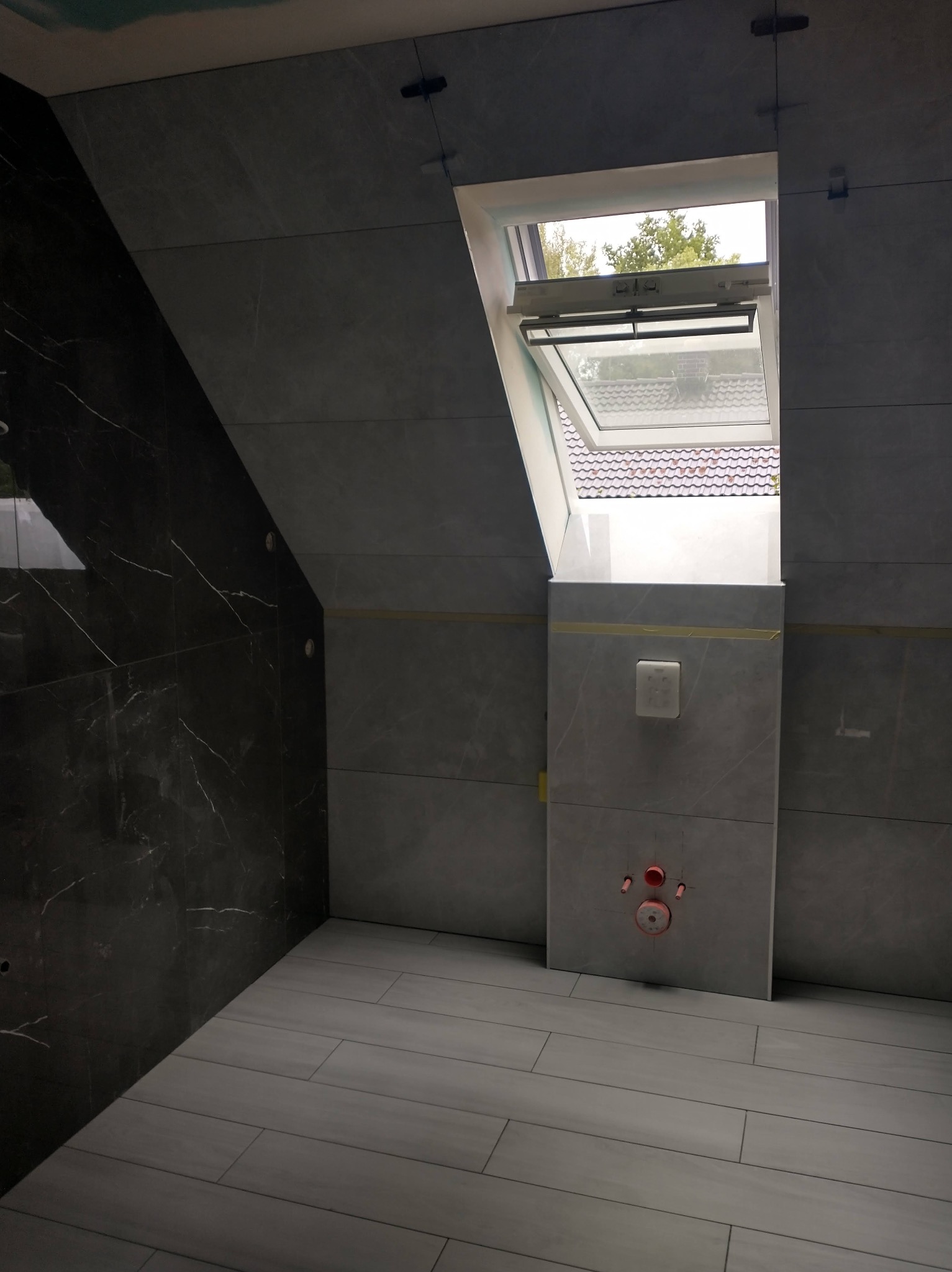 Łazienka na poddaszu w trakcie wykańczania: szare płytki imitujące kamień na ścianach i podłodze, okno dachowe z widokiem na drzewo, fragment ściany z czarną marmurową okładziną, widoczne elementy...