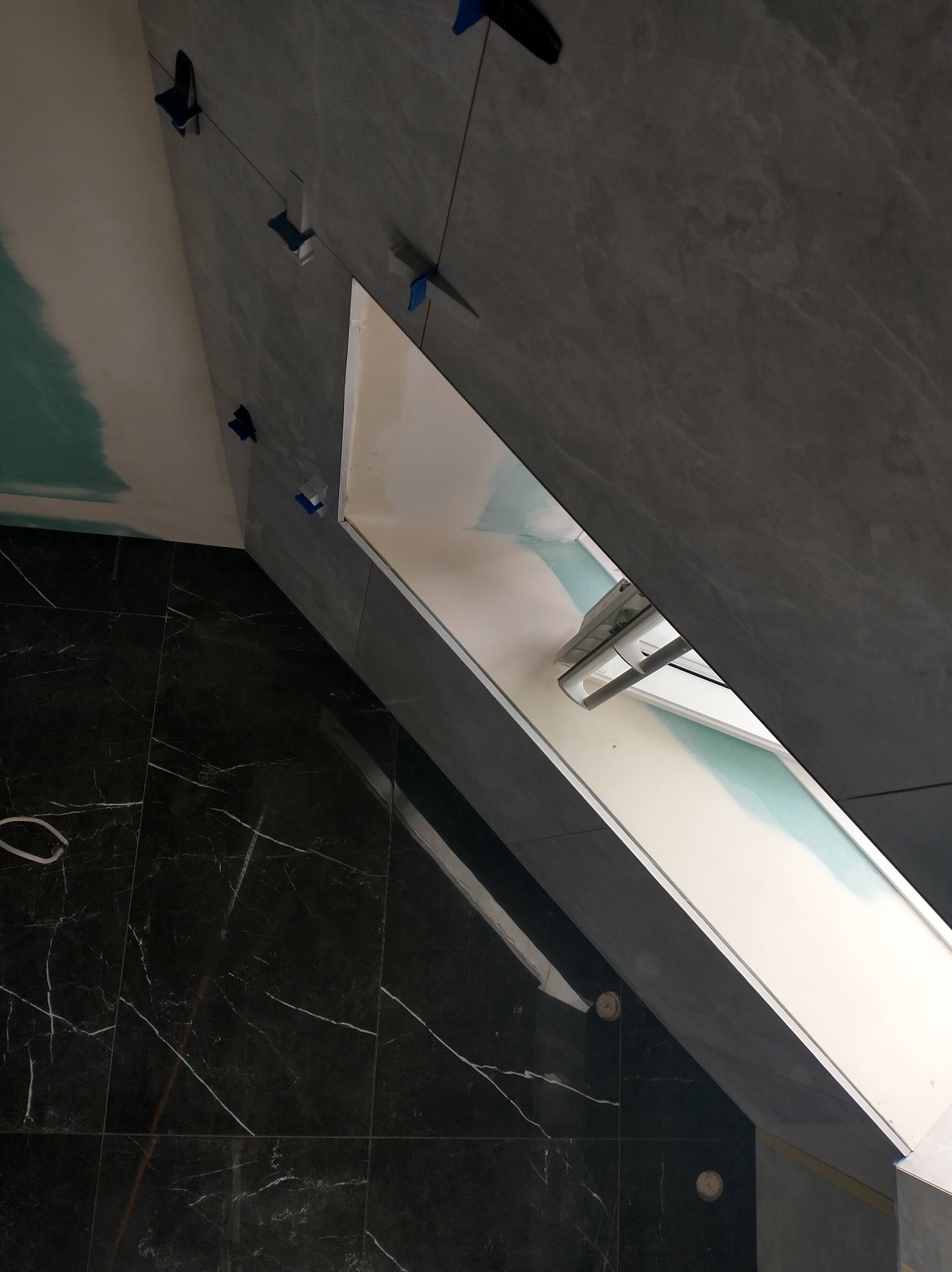 Montaż szarych płytek na ścianie w łazience, widoczne niebieskie klipsy poziomujące, czarna marmurowa podłoga i biała zabudowa wnęki.