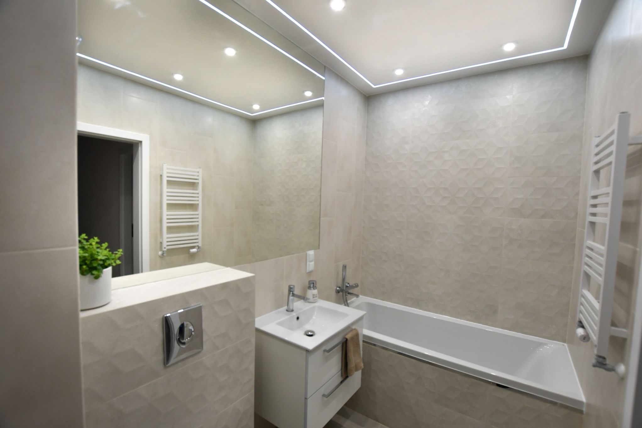 Jasna łazienka z geometrycznymi płytkami, podświetlanym lustrem, białą umywalką z szafką, grzejnikiem drabinkowym i wanną.