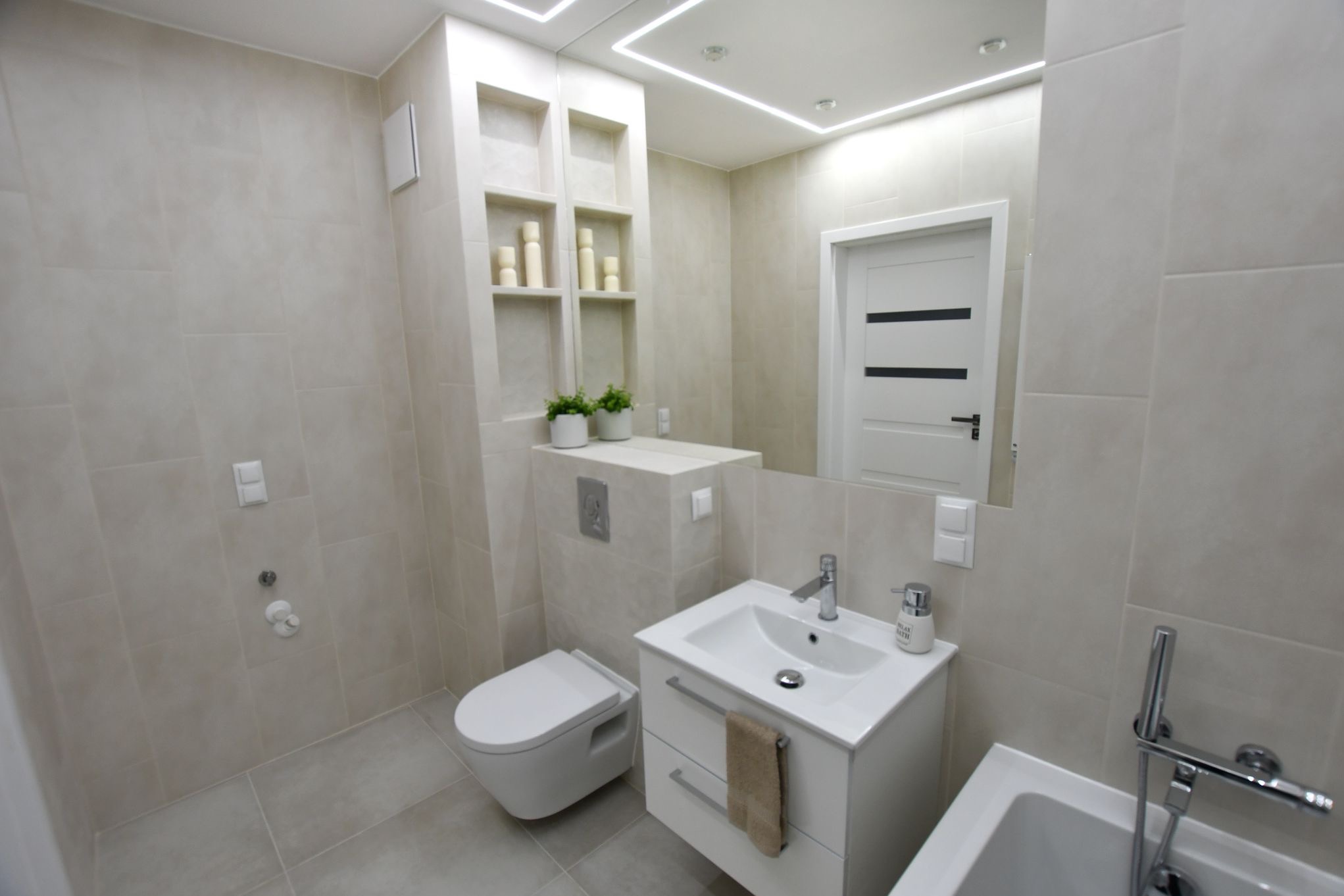 Jasna łazienka z beżowymi płytkami, podwieszana toaleta, biała szafka pod umywalką z szufladami i ręcznikiem, lustro z oświetleniem LED, oraz wnęka z półkami i dekoracjami.