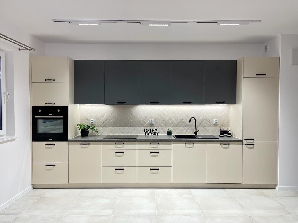 Nowoczesna kuchnia z beżowymi i grafitowymi szafkami, podświetleniem LED pod górnymi szafkami, płytkami ułożonymi w jodełkę na ścianie oraz czarną armaturą i napisem dekoracyjnym 'Dzień Dobry'.