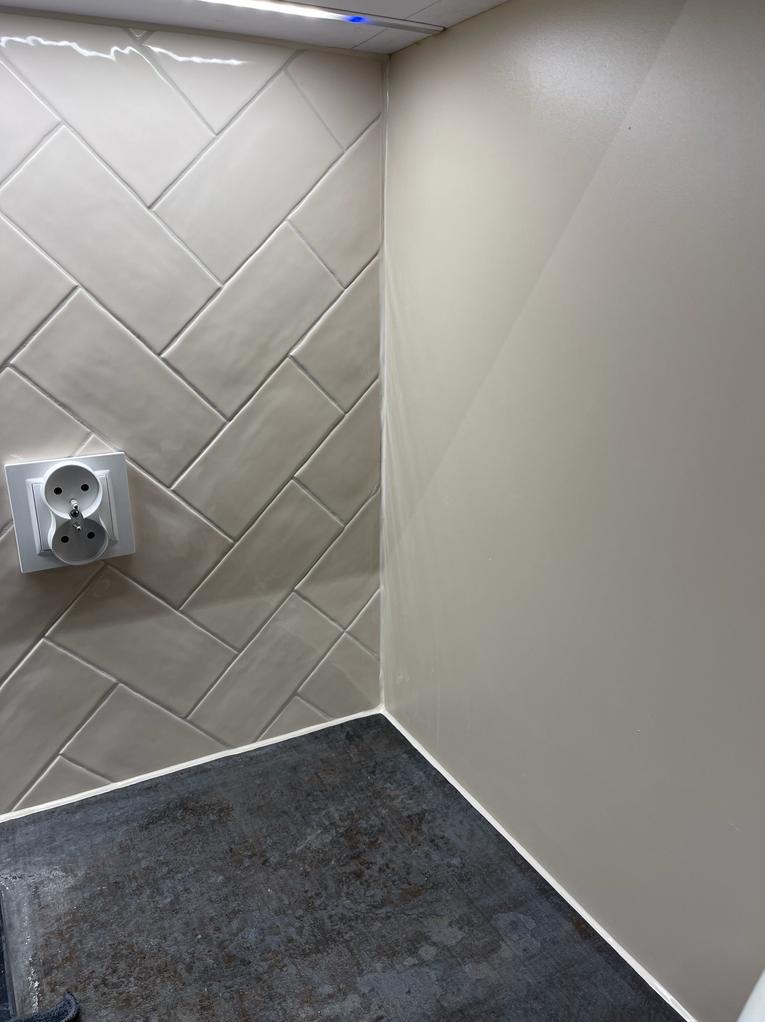 Ściana wyłożona szarymi płytkami ułożonymi w jodełkę z gniazdkiem elektrycznym, połączona z gładką, pomalowaną na szaro ścianą, widoczna szara podłoga