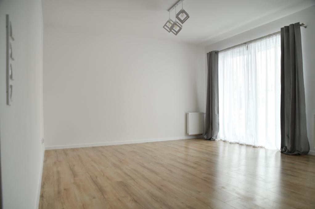 Jasne, puste pomieszczenie z panelami podłogowymi imitującymi drewno, białymi ścianami, szarymi zasłonami, białym grzejnikiem i nowoczesną lampą sufitową.