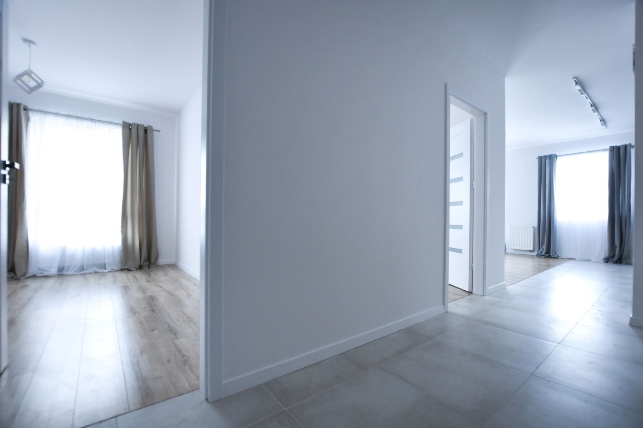 Jasne wnętrze mieszkania z szarymi płytkami podłogowymi i drewnianą podłogą w pokoju obok, widoczne przez otwarte drzwi, z szarymi zasłonami i białymi ścianami.