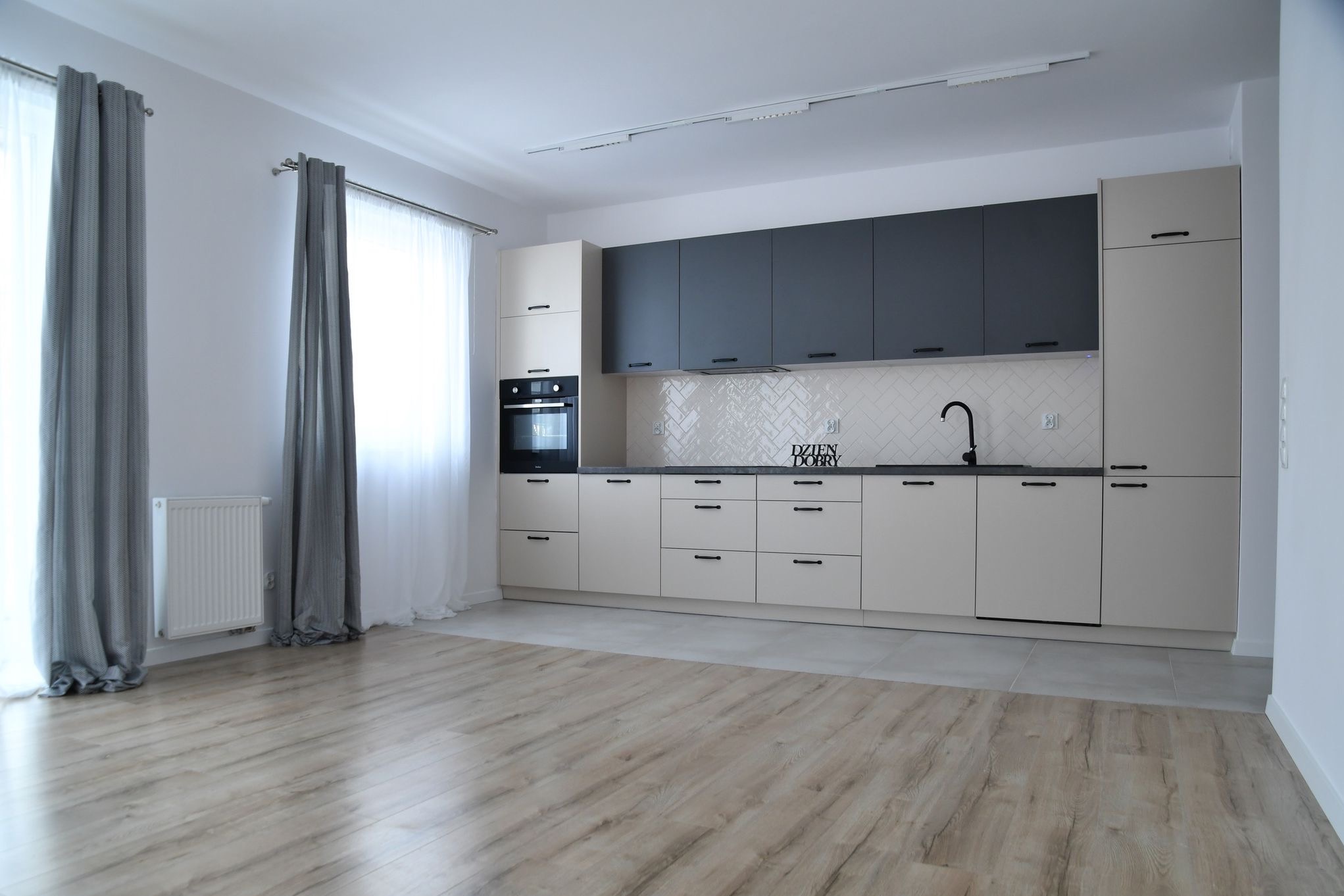 Jasna kuchnia z szarymi górnymi szafkami, białymi płytkami ułożonymi w jodełkę i drewnianą podłogą. Na ścianie napis 'Dzień Dobry'.