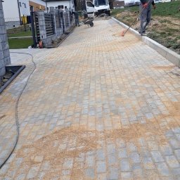 Wojciech SIkora - Ułożona szara kostka brukowa, zasypywana piaskiem, z widocznym fragmentem ogrodzenia z szarych elementów betonowych i pracownikiem wygładzającym powierzchnię miotłą.