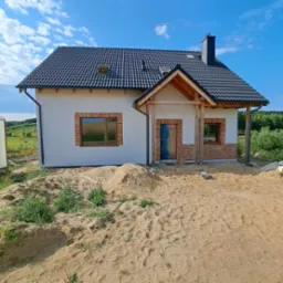 Nowo wybudowany dom jednorodzinny z białymi ścianami, czarnym dachem i ceglaną dekoracją wokół okien, otoczony piaskiem i zielenią, w słoneczny dzień.