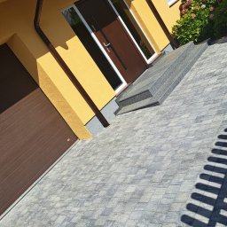 Wojciech SIkora - Żółty dom jednorodzinny z brązowymi drzwiami i bramą garażową, szara kostka brukowa na podjeździe, schody wejściowe z szarego kamienia, krzewy hortensji.