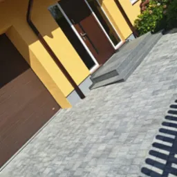 Żółty dom jednorodzinny z brązowymi drzwiami i bramą garażową, szara kostka brukowa na podjeździe, schody wejściowe z szarego kamienia, krzewy hortensji.