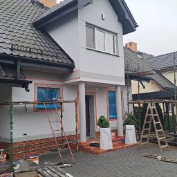 Wojciech SIkora - Dom w trakcie prac elewacyjnych: szara fasada z pomarańczowymi akcentami wokół okien i kolumn, okna zabezpieczone folią, rusztowania, kostka brukowa na podjeździe.