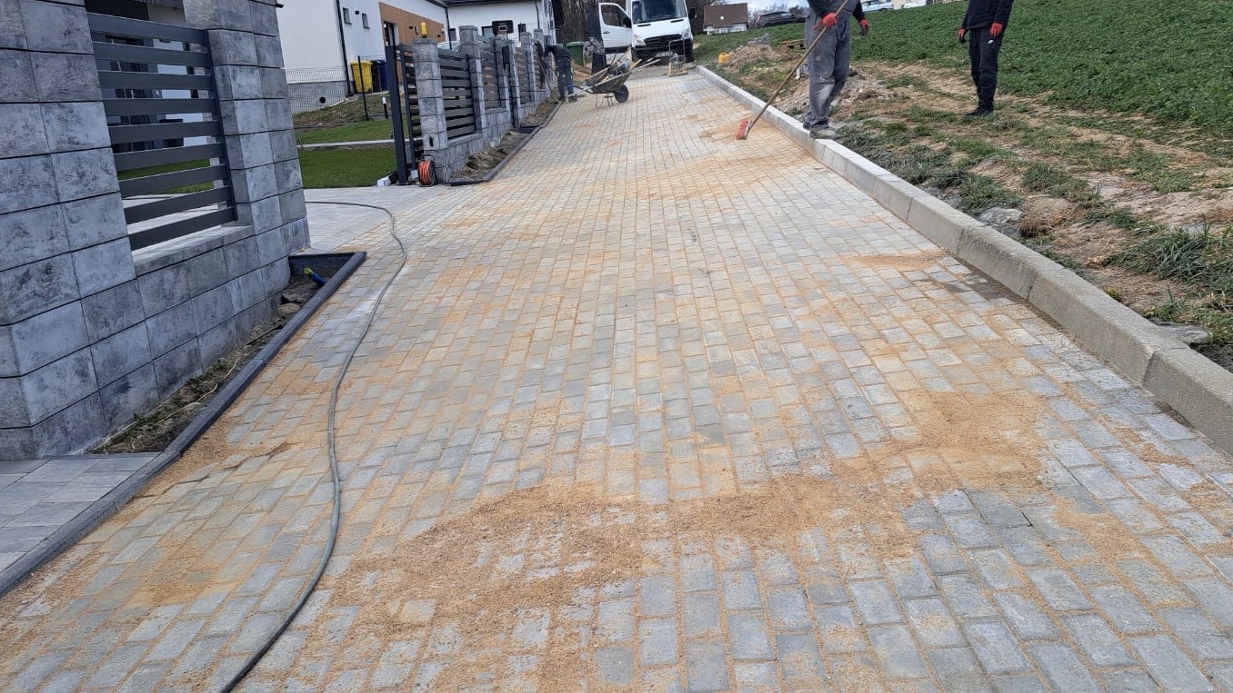 Ułożona szara kostka brukowa, zasypywana piaskiem, z widocznym fragmentem ogrodzenia z szarych elementów betonowych i pracownikiem wygładzającym powierzchnię miotłą.